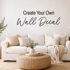 May include: A black wall decal featuring the text Create Your Own Wall Decal in a combination of sans-serif and script fonts. The text is applied to a plain white wall above a white sofa with beige throw pillows and a woven ottoman.