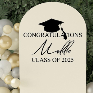 May include: A white graduation sign with black text that reads "Congratulations Maddie Class of 2025". A black graduation cap silhouette is above the text.