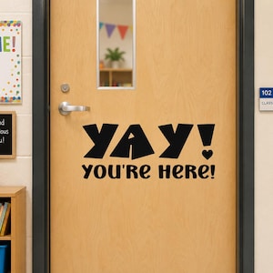 May include: A light wood classroom door featuring a black vinyl decal that reads YAY! YOU'RE HERE! in a bold, stylized font. The door includes a silver metal handle, a lock, and a rectangular glass window panel centered in the upper portion.
