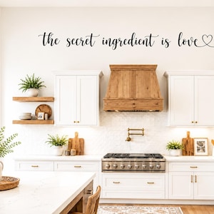 May include: A black wall decal featuring the cursive script text "the secret ingredient is love" followed by a small heart outline. The lettering is applied to a white wall above a kitchen range hood, white cabinetry, and wooden shelving.