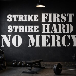 May include: A black wall featuring large, white, stencil-style lettering that reads STRIKE FIRST, STRIKE HARD, NO MERCY. The wall is situated in a gym environment containing a weight rack with dumbbells, a flat exercise bench, and a barbell.
