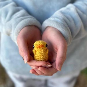 May include: A small yellow crocheted chick with black eyes and an orange beak is being held in the palms of two hands.