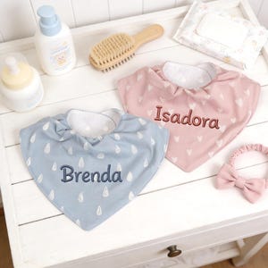 May include: Two personalized baby bibs, one light blue with raindrop design and the name "Brenda", the other pink with hearts and the name "Isadora". A matching pink bow headband is also present.