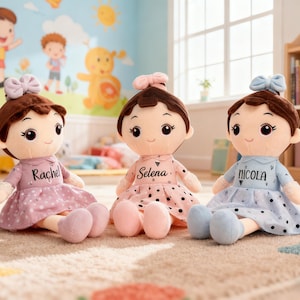 May include: Three plush dolls with brown hair and bows, wearing dresses in pink, light blue, and pink. Each doll has a name on its dress: Rachel, Selena, and Nicola. The dolls are sitting on a patterned rug.