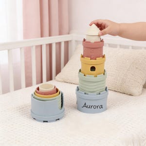 May include: Stacking toy set with castle-shaped cups in pastel colors. The set includes a tower of cups with the name "Aurora" on the base, and a smaller set of nested cups. The tower is being assembled by a child.
