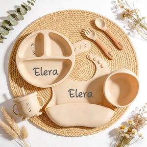 Könnte beinhalten: Ein beiges Baby-Ess-Set mit Giraffen-Design, bestehend aus einem geteilten Teller, einer Schüssel, einer Lätzchen, einem Becher, einer Gabel und einem Löffel. Der Name "Elera" ist auf Teller, Lätzchen und Löffel aufgedruckt. Das Set ist auf einer gewebten Matte angeordnet.