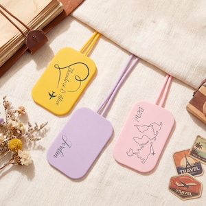 May include: Three luggage tags in yellow, lavender, and pink. The yellow tag has the text "Theodore & Alice" and an airplane graphic. The lavender tag says "Jordan". The pink tag has a map graphic and the text "BeW".