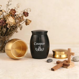 Puede incluir: Una urna negra para mascotas con el nombre "Cooper" y un diseño de huellas de patas. Una urna dorada está abierta al lado. También hay una pequeña cruz de madera y una tapa dorada, junto con un pequeño ramo de flores secas.