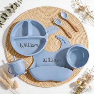 Puede incluir: Juego de alimentación para bebés en azul claro, con plato dividido, cuenco, babero, taza, tenedor y cuchara. Cada artículo está personalizado con el nombre "William" y una ilustración de león. Cuchara y tenedor con mango de madera.
