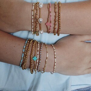May include: An assortment of beaded bracelets in gold, white, pink, and turquoise. Some bracelets feature pearls and cross charms. The bracelets are displayed on wrists against a light blue background.