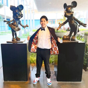 May include: A young person wearing a black suit jacket with a colorful comic book print lining, a white dress shirt, a black bow tie, and black pants. The person is standing between two bronze statues of Mickey Mouse and Minnie Mouse.