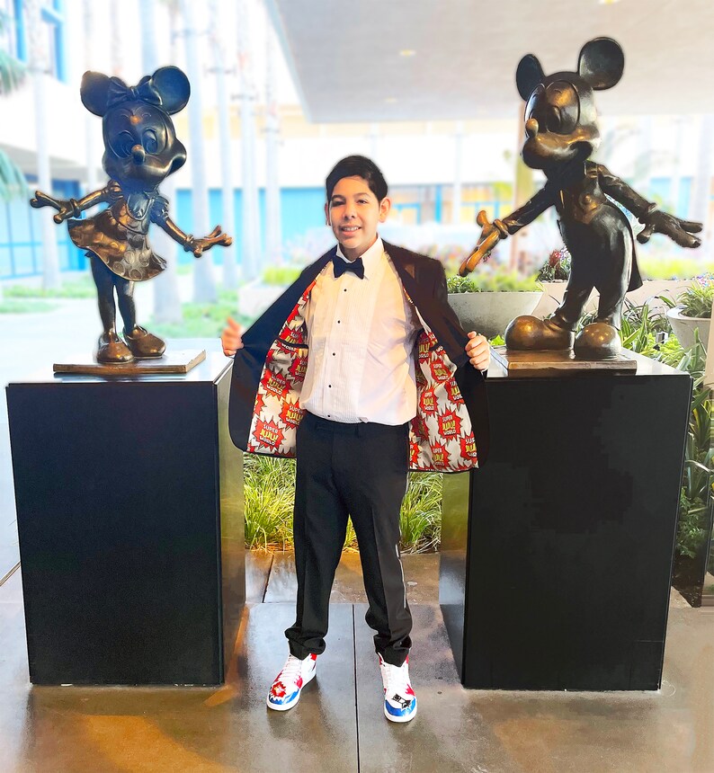 May include: A young person wearing a black suit jacket with a colorful comic book print lining, a white dress shirt, a black bow tie, and black pants. The person is standing between two bronze statues of Mickey Mouse and Minnie Mouse.