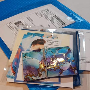 May include: A blue and gold abstract art print with a silver ribbon tied around it. The print is folded in half and is sitting on top of a stack of white envelopes and a blue and white patterned fabric. The text "Designer Scarves by Marlena.com" is visible on one of the envelopes.