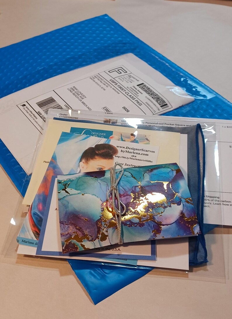 May include: A blue and gold abstract art print with a silver ribbon tied around it. The print is folded in half and is sitting on top of a stack of white envelopes and a blue and white patterned fabric. The text "Designer Scarves by Marlena.com" is visible on one of the envelopes.