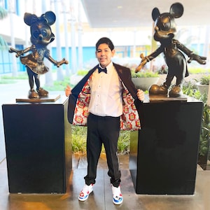 May include: A young person wearing a black suit jacket with a colourful comic book print lining, a white dress shirt, a black bow tie, and black trousers. The person is standing between two bronze statues of Mickey Mouse and Minnie Mouse.