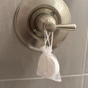 Puede incluir: Un limpiador de cabezal de ducha blanco en una bolsa de malla blanca colgando de una manija de ducha plateada con la etiqueta "DELTA".