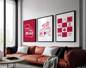 May include: Three framed prints with red and white designs hang above a leather sofa. The prints feature a Georgia outline with text, a bulldog illustration, and a checkerboard pattern with text and symbols. The room has a modern design.