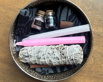 May include: A metal bowl with a black and white patterned cloth, two small glass jars labeled "Rose Quartz" and "Amethyst", a white candle, a pink candle, and a bundle of sage.