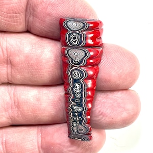 以下が含まれることがあります： 鮮やかな赤色の外観と、黒、白、青の複雑な模様が特徴的な、手作りの細長いビーズ。ビーズのデザインは同心円と流れるようなラインが特徴で、ユニークな美しさを生み出しています。ビーズは手で持たれています。