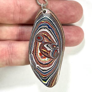 Könnte beinhalten: Ein ovaler Anhänger mit einem auffälligen Schichtmuster in Rot-, Blau-, Gelb- und Weißtönen, der an einer Silberkette hängt. Das Design ähnelt einem Stein oder Mineral mit konzentrischen Ringen. Das Muster des Anhängers ist auffällig.