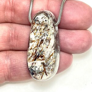May include: A clear, oval pendant with a silver chain. The pendant features a mix of white, brown, and gray inclusions, creating a unique, natural pattern. The chain is a delicate, snake-style design, adding a touch of elegance to the necklace.