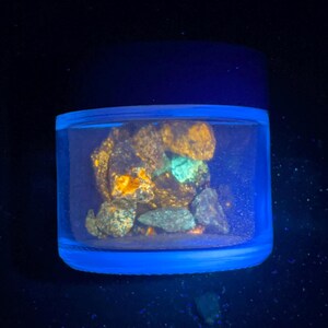 May include: A clear, cylindrical jar with a dark blue lid, illuminated by a blue light. Inside, various rocks and minerals glow with orange, green, and gray hues, creating a striking contrast against the dark background.