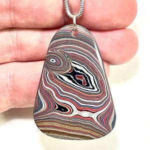 Könnte beinhalten: Ein tropfenförmiger Anhänger mit einem auffälligen Muster aus roten, schwarzen, weißen und braunen Schichten. Der Anhänger hängt an einer silbernen Kette. Das Design des Steins ähnelt einem farbenfrohen, abstrakten Auge.