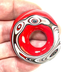 以下が含まれることがあります： 中央に穴のある赤い円形のペンダント。ペンダントは、赤い背景に対して、白、黒、グレーの渦巻き模様が特徴です。デザインは石やユニークな芸術作品を連想させます。