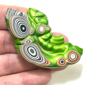 以下が含まれることがあります： メタリック仕上げの、手作りの緑色の葉の形をしたペンダント。ペンダントは、黒、白、グレーの同心円とオレンジ色のアクセントが特徴です。ユニークなデザインが手で持たれています。