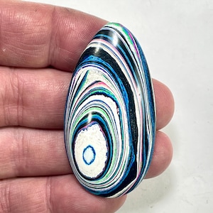 May include: An oval-shaped stone with a swirling pattern of black, white, blue, and green. The stone has a polished surface and a unique, eye-like design in the center. The stone is held in a hand.