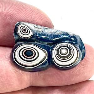 以下が含まれることがあります： 黒、白、ピンクの印象的な同心円模様が施された、磨かれた濃い青色の石。 石の表面は滑らかで光沢があり、独特の目のように見えます。 石は指の間に挟まれています。