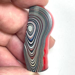 以下が含まれることがあります： 黒、白、青、赤の渦巻き模様が特徴的な、層状の石のオブジェ。手で持ち、そのユニークでカラフルなデザインを披露しています。ベースは無地の赤色です。