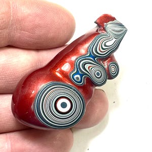 以下が含まれることがあります： 光沢のある仕上がりの赤い涙滴型のオブジェクト。表面には、黒、白、青、赤の同心円と渦巻き模様のユニークなパターンがあります。オブジェクトは手で持たれています。