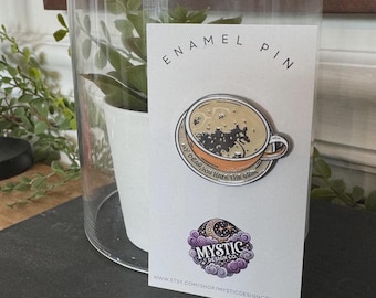 May include: An enamel pin featuring a coffee cup design with the text "MY DEAR, YOU HAVE THE GRIM". The pin is on a white card with the text "ENAMEL PIN" and the Mystic Design Co. logo. The pin is displayed under a glass cloche.