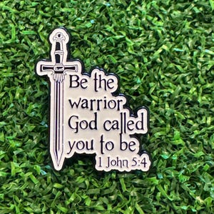May include: A white and black enamel pin featuring a sword and the text "Be the warrior God called you to be 1 John 5:4". The pin is set against a green, textured background.