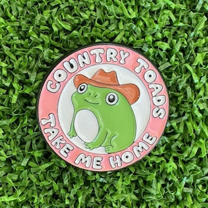 May include: A round pin featuring a green frog wearing a brown cowboy hat. The pin has a pink border with the words "COUNTRY TOADS TAKE ME HOME" in white lettering. The frog has large, round eyes.
