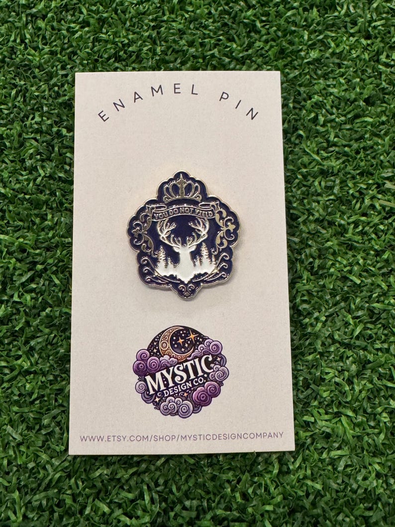 May include: An enamel pin on a white card. The pin features a deer with the text "YOU DO NOT YIELD" in a navy blue and white design. Below is a purple and white logo with the text "MYSTIC DESIGN CO." and a website address.
