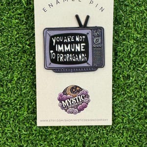 May include: An enamel pin featuring a vintage television design. The screen displays the text "YOU ARE NOT IMMUNE TO PROPAGANDA" in white. The pin is on a cream-colored card with the words "ENAMEL PIN" at the top and a logo at the bottom.