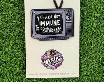 May include: An enamel pin featuring a vintage television design. The screen displays the text "YOU ARE NOT IMMUNE TO PROPAGANDA" in white. The pin is on a cream-colored card with the words "ENAMEL PIN" at the top and a logo at the bottom.