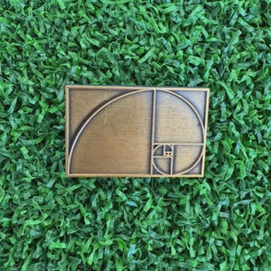 May include: A rectangular, bronze-colored metal pin featuring the golden ratio spiral design. The pin is set against a backdrop of artificial green grass.