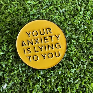 May include: A round, yellow enamel pin with the phrase "YOUR ANXIETY IS LYING TO YOU" in black lettering. The pin has a black border and is set against a background of green artificial turf.