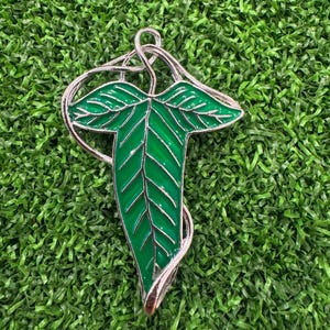 May include: A silver-toned pendant in the shape of a stylized green leaf with a detailed vein pattern. The leaf is outlined in silver and has a glossy finish. The pendant is set against a green background.