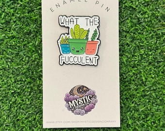 May include: An enamel pin featuring three potted succulents with smiling faces. The pin has the text "WHAT THE FUCCULENT" in a playful font. The pin is on a light-colored card with the text "ENAMEL PIN" at the top.