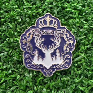May include: A decorative pin with a blue and gold design. The pin features a white stag silhouette, trees, and the text "YOU DO NOT YIELD" on a banner. The pin has a crown at the top and is set against a green background.