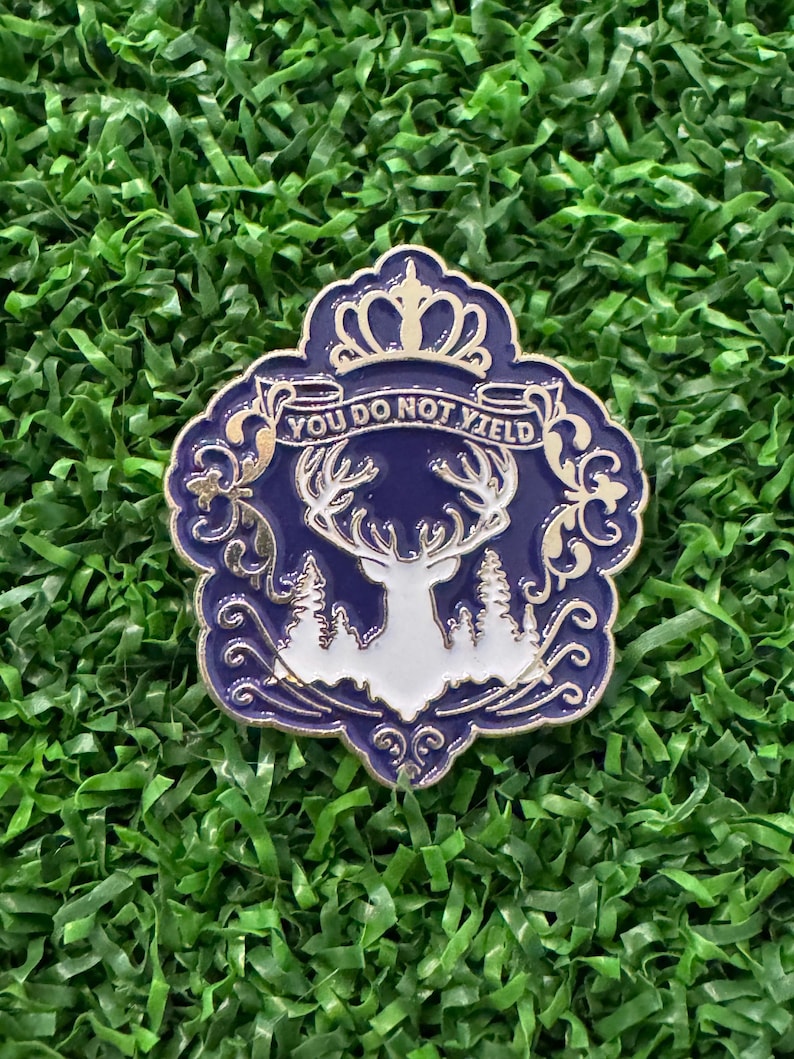 May include: A decorative pin with a blue and gold design. The pin features a white stag silhouette, trees, and the text "YOU DO NOT YIELD" on a banner. The pin has a crown at the top and is set against a green background.