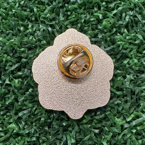 May include: A gold-colored metal pin with a flower shape. The pin has a textured surface and a gold-colored clasp on the back. The pin is set against a green background.