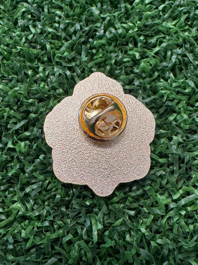 May include: A gold-colored metal pin with a flower shape. The pin has a textured surface and a gold-colored clasp on the back. The pin is set against a green background.