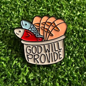 May include: A metal enamel pin featuring a white basket containing two fish, one light blue and one red, alongside several tan loaves of bread. The basket is inscribed with the black text "GOD WILL PROVIDE" in a bold, sans-serif font.