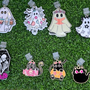 May include: A collection of Halloween-themed acrylic charms featuring ghosts and black cats. The charms include ghosts with boba tea, flowers, and reading a book. Other charms include black cats with pink accents and a coffin with skeletons.