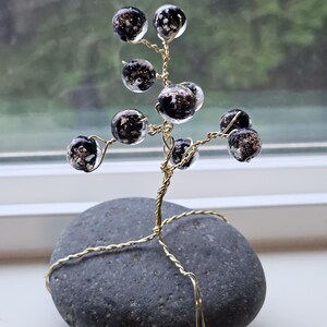 Könnte beinhalten: Eine Drahtskulptur eines Baumes mit schwarzen und goldenen Glasperlen, die auf einem grauen Steinsockel steht.